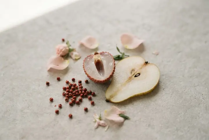 A halved lychee, a slice of pear, pink peppercorns and loose rose petals arranged on pale stone