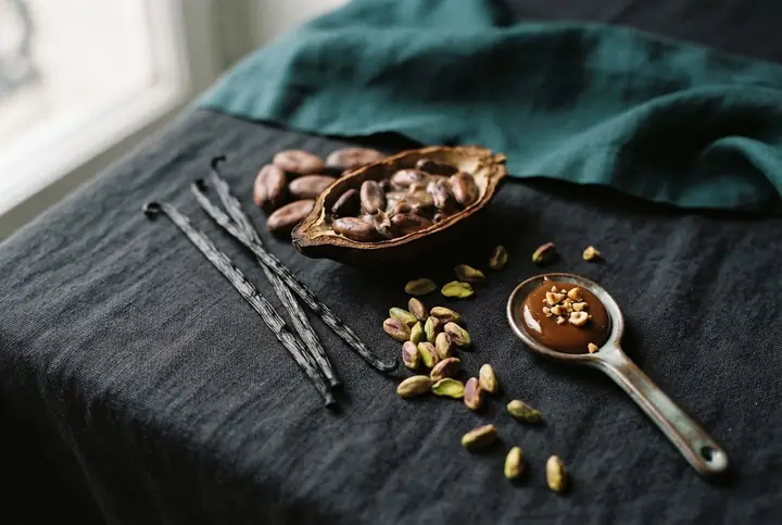 Vanilla pods, a split cocoa pod, shelled pistachios and a spoon of praline on dark linen