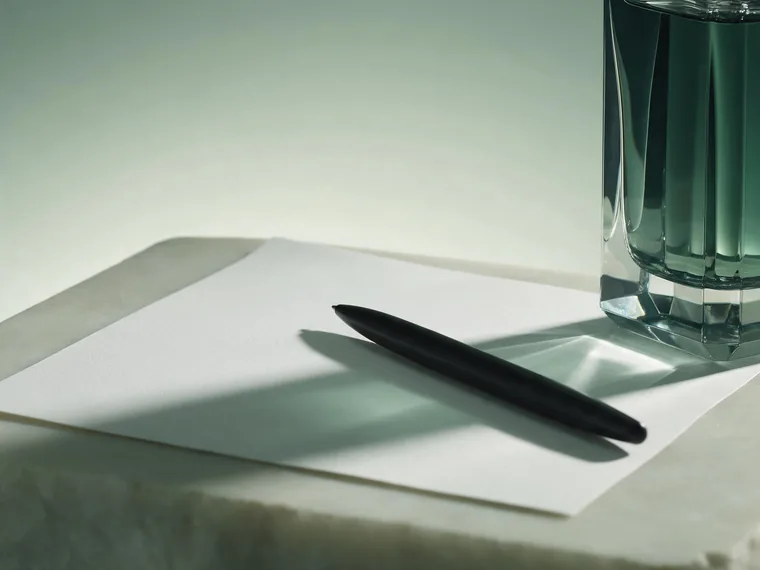 A blank sheet of white paper with a matte black pen resting on it, beside the base of a faceted crystal perfume bottle